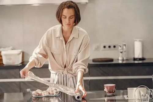 Image is of a young woman in the kitchen measuring out aluminum foil to cover a dish with. 